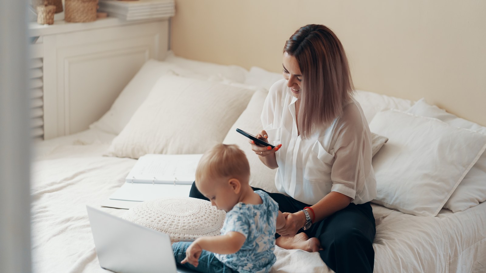 Mother checking baby monitor video stream on smartphone while infant rests peacefully in travel crib