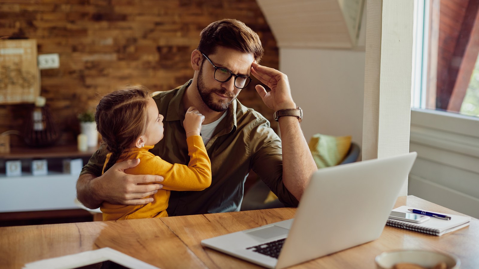 A focused father working on his laptop in a modern home office while monitoring his sleeping baby peacefully in the background.