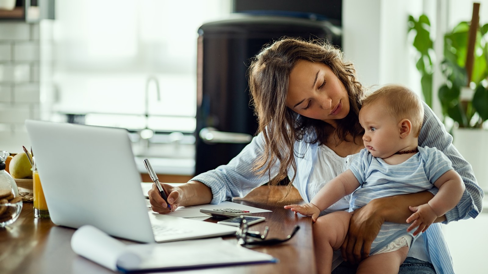 A working mother monitoring her sleeping baby on a laptop computer screen via a floating Picture-in-Picture window while working at her home office desk.