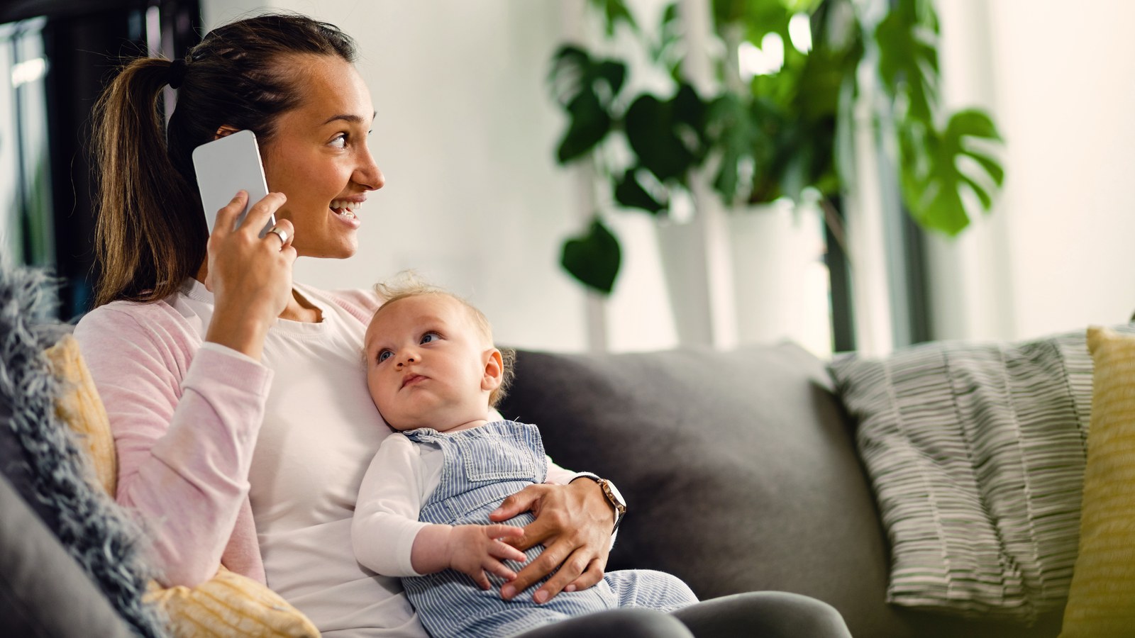 A happy mother relaxing in her living room speaking softly into her phone to soothe her sleeping baby in the nursery.